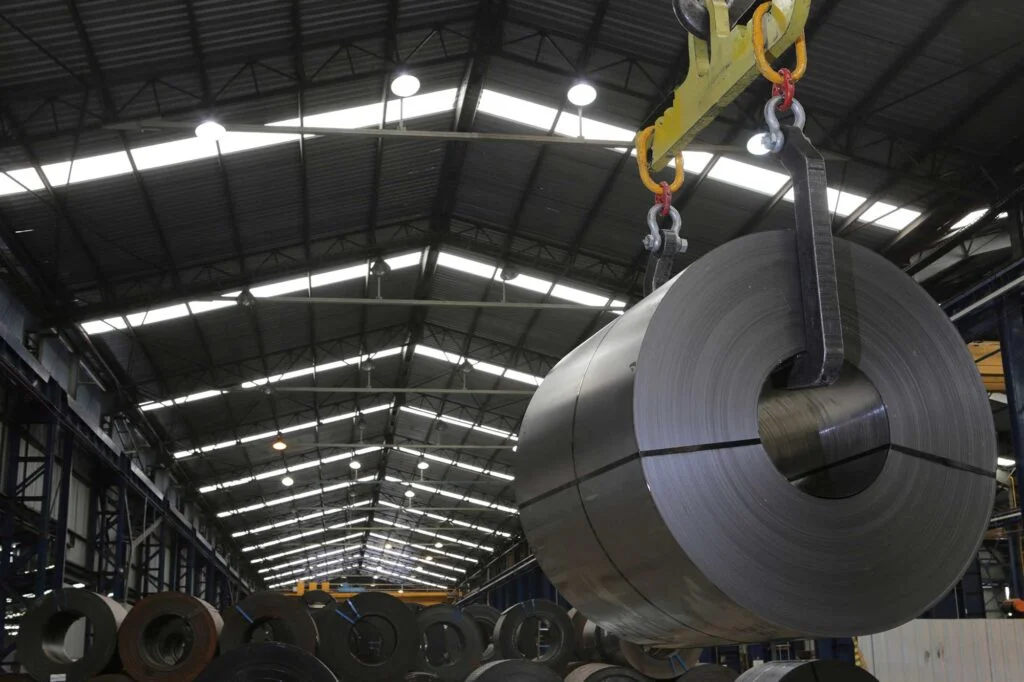 Bobina de acero suspendida por una grúa puente dentro de una planta sider?rgica, con otras bobinas met?licas apiladas en el interior de una nave industrial.