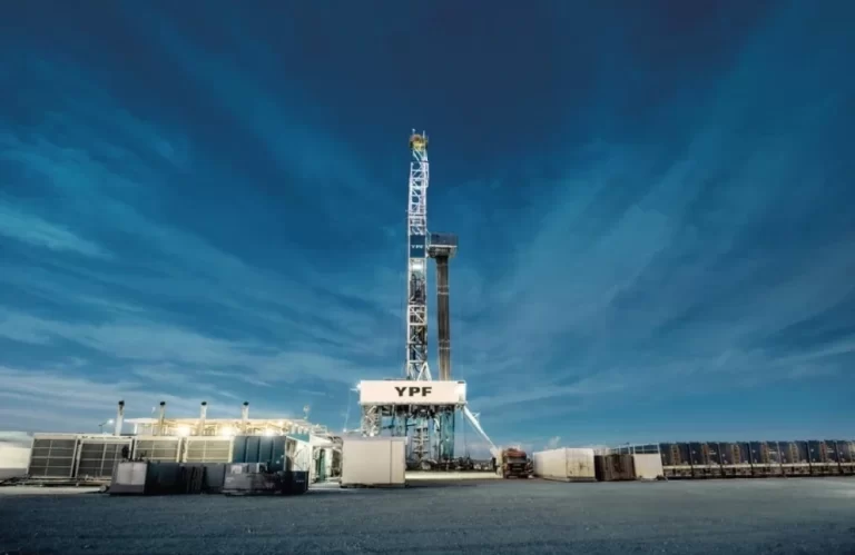 Planta de perforación petrolera de YPF en una zona industrial a cielo abierto, con una torre de extracción en el centro, contenedores y equipos t?cnicos alrededor, bajo un cielo azul intenso con nubes alargadas.