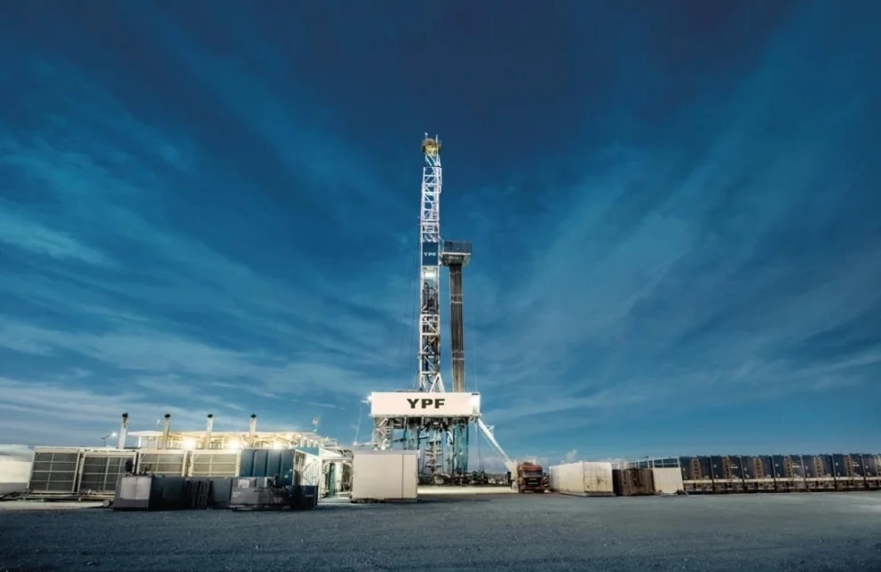 Planta de perforación petrolera de YPF en una zona industrial a cielo abierto, con una torre de extracción en el centro, contenedores y equipos t?cnicos alrededor, bajo un cielo azul intenso con nubes alargadas.