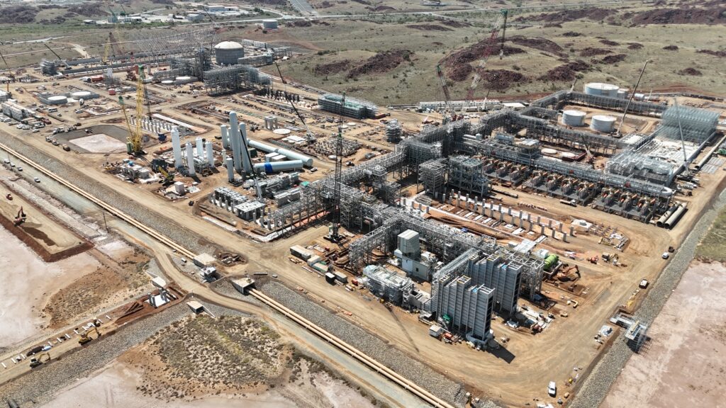 Instalación industrial en construcción con múltiples estructuras met?licas, equipos de proceso y grúas en un paisaje seco.