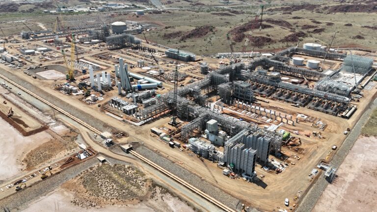 Instalación industrial en construcción con múltiples estructuras metálicas, equipos de proceso y grúas en un paisaje seco.