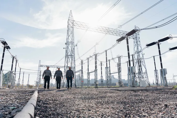 Tres operarios caminan dentro de una subestación eléctrica rodeados de torres, cables y equipos de alta tensión.