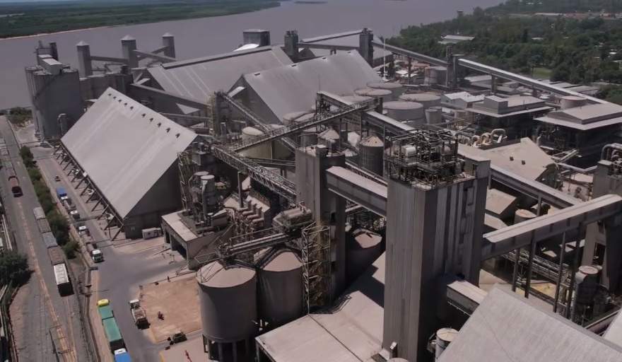 Vista aérea de una planta industrial de procesamiento junto a un río, con silos, cintas transportadoras y grandes estructuras fabriles.