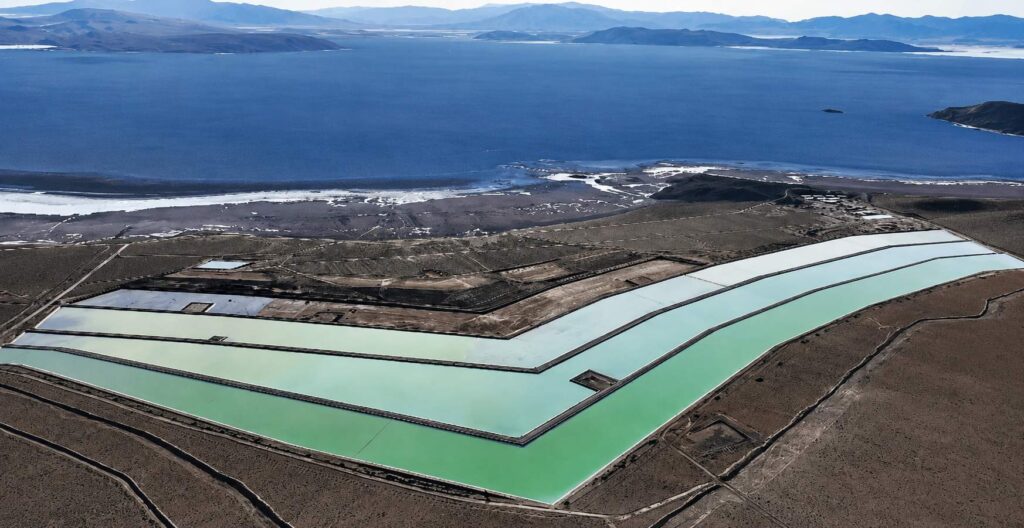 Vista aérea de piscinas de evaporación utilizadas para la producción de litio, ubicadas en una zona árida junto a un gran cuerpo de agua rodeado de montañas.