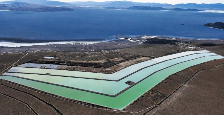 Vista aérea de piscinas de evaporación utilizadas para la producción de litio, ubicadas en una zona árida junto a un gran cuerpo de agua rodeado de montañas.