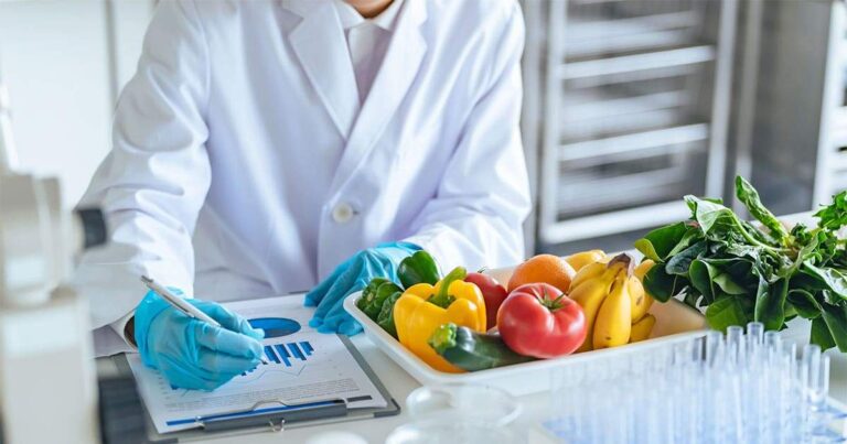 Profesional de laboratorio analiza datos junto a una bandeja con frutas y verduras frescas, en un entorno de control de calidad y seguridad alimentaria.