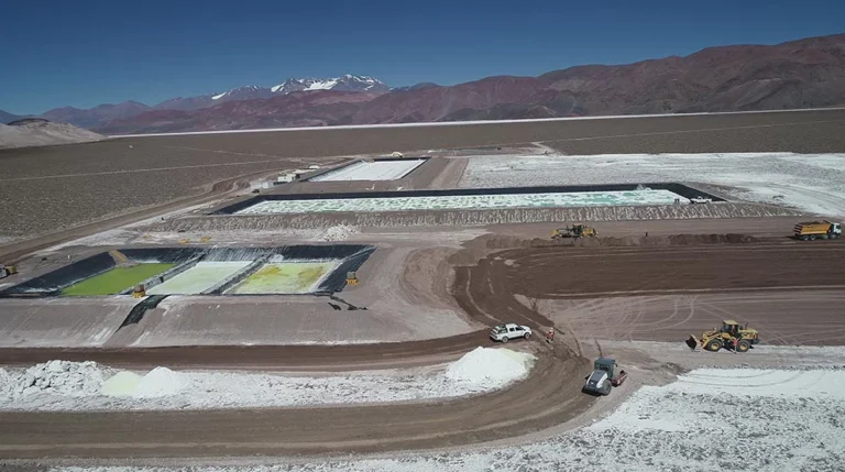 Vista panorámica de un proyecto de extracción de litio en un salar de alta montaña, con piletas de evaporación de distintos colores, maquinaria vial, caminos internos y la cordillera al fondo.