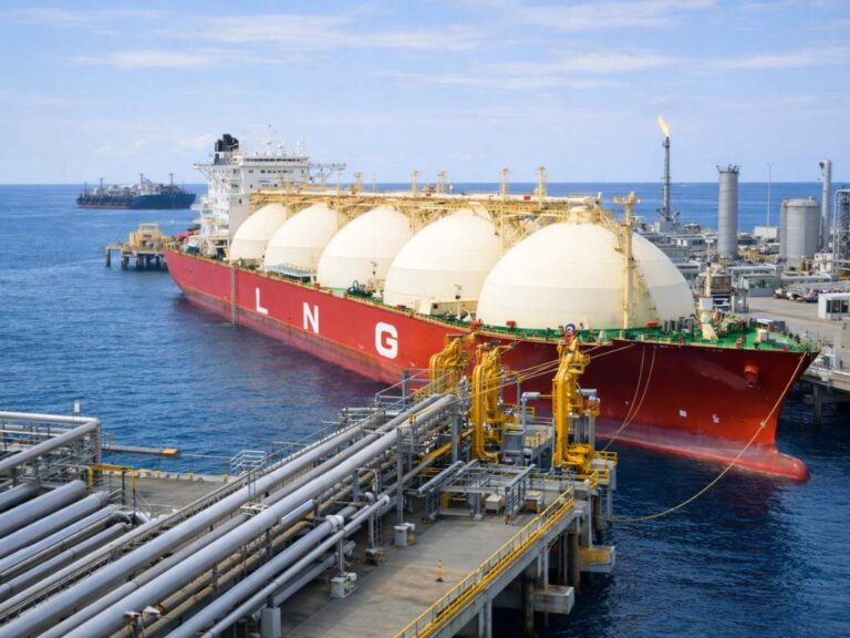 Buque metanero atracado en una terminal portuaria de gas natural licuado (GNL), conectado a la infraestructura de carga y descarga mediante tuberías industriales, con instalaciones energéticas y otro barco al fondo.