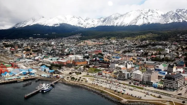 Vista aérea de una ciudad costera con puerto, edificios urbanos y la cordillera nevada al fondo.