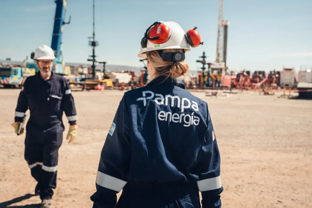 Trabajadora de Pampa Energía en un yacimiento petrolero, con equipos de perforación e infraestructura industrial al fondo.