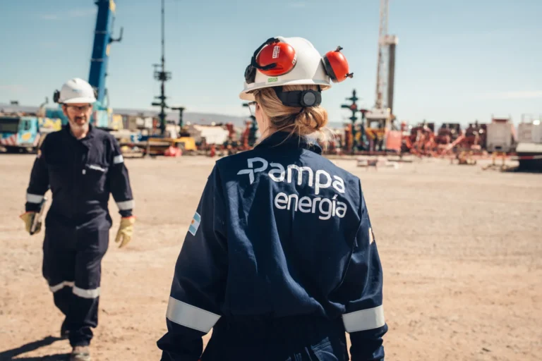 Trabajadora de Pampa Energía en un yacimiento petrolero, con equipos de perforación e infraestructura industrial al fondo.