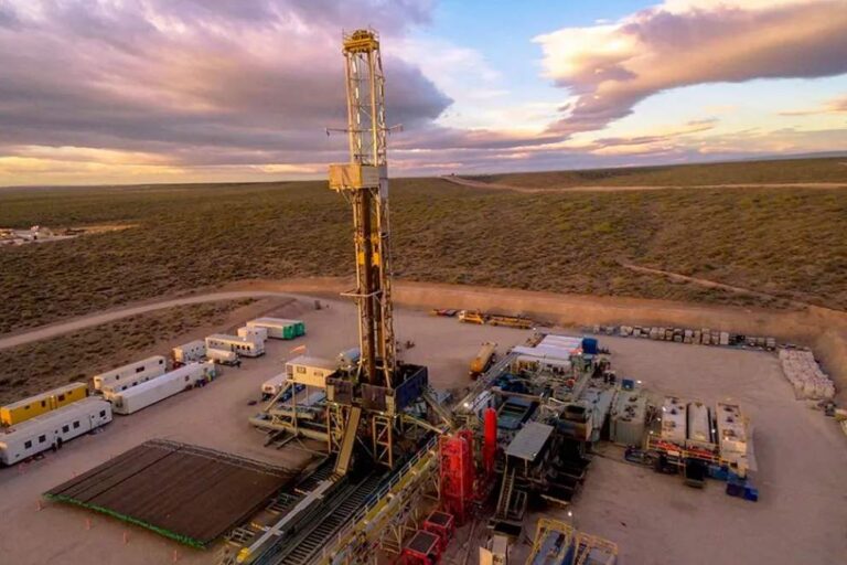 Vista aérea de un equipo de perforación petrolera en un yacimiento de Vaca Muerta, con plataformas, módulos operativos, equipos auxiliares y caminos de acceso, en un entorno de estepa patagónica durante el atardecer.