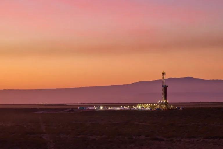 Equipo de perforación petrolera iluminado al atardecer en un yacimiento terrestre, con un paisaje árido y montañas en el horizonte.
