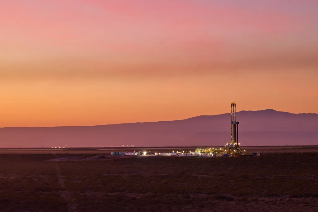 Equipo de perforación petrolera iluminado al atardecer en un yacimiento terrestre, con un paisaje árido y montañas en el horizonte.