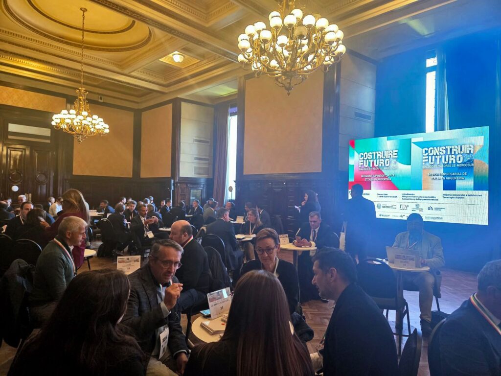 Encuentro empresarial en un salón, con participantes reunidos en mesas de trabajo y una pantalla que presenta el evento Construire Futuro, sobre oportunidades de negocios entre Italia y Argentina.