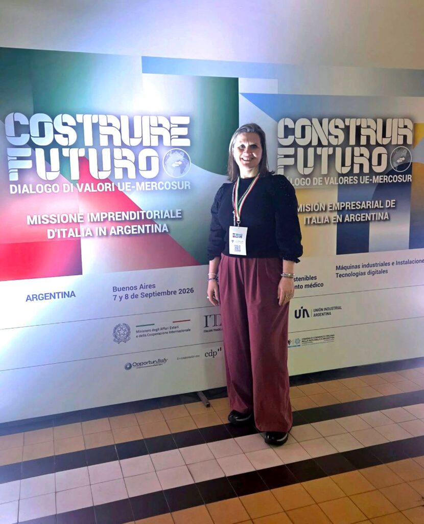 Imagen de una mujer con una credencial de prensa fotografiada delante de un pizarrón que anuncia un evento llamado "Contruire Futuro: Dialogo di valori UE-Mercosur - Missione Imprenditoriale D'Italia in Argentina".